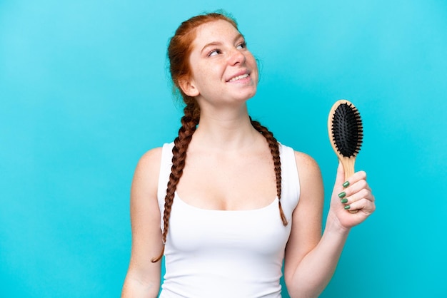Junge kaukasische rötliche Frau mit Haarkamm isoliert auf blauem Hintergrund, die lächelnd aufblickt