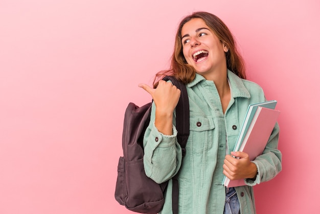 Junge kaukasische Studentenfrau, die Bücher einzeln auf rosafarbenem Hintergrund hält, zeigt mit dem Daumenfinger weg, lacht und sorglos.