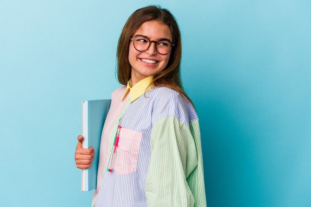 Junge kaukasische Studentin, die Bücher auf blauem Hintergrund hält, sieht beiseite lächelnd, fröhlich und angenehm aus.