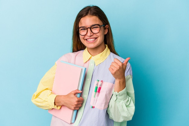 Junge kaukasische Studentin, die Bücher auf blauem Hintergrund isoliert hält und lächelt und Daumen hochhebt