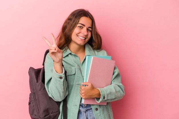 Junge kaukasische Studentin, die Bücher isoliert auf rosa Hintergrund hält und Nummer zwei mit den Fingern zeigt.