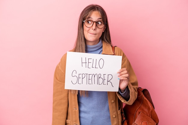 Junge kaukasische Studentin, die ein hallo September-Plakat einzeln auf rosafarbenem Hintergrund hält, verwirrt, fühlt sich zweifelhaft und unsicher.