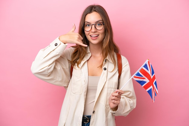 Junge kaukasische Studentin, die eine Flagge des Vereinigten Königreichs hält, die auf rosa Hintergrund isoliert ist und eine Telefongeste macht Ruf mich zurück