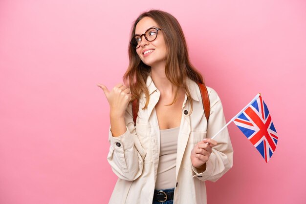 Junge kaukasische Studentin, die eine Flagge des Vereinigten Königreichs isoliert auf rosafarbenem Hintergrund hält, die zur Seite zeigt, um ein Produkt zu präsentieren