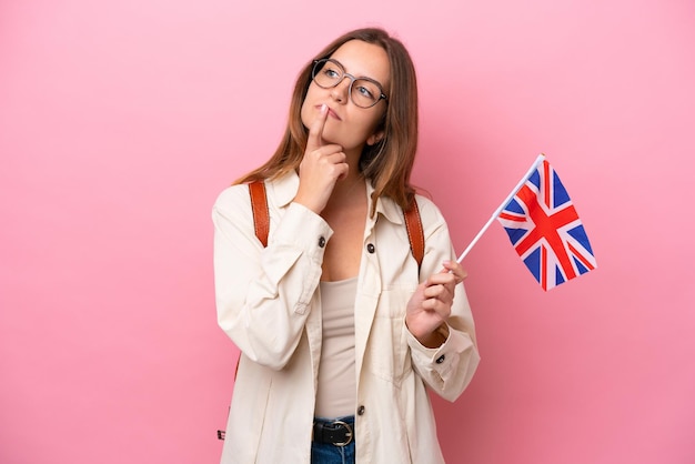 Junge kaukasische Studentin, die eine Flagge des Vereinigten Königreichs isoliert auf rosafarbenem Hintergrund hält und beim Aufblicken Zweifel hat