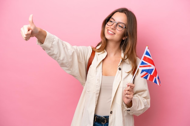Junge kaukasische Studentin, die eine Flagge des Vereinigten Königreichs isoliert auf rosafarbenem Hintergrund hält und eine Geste mit dem Daumen nach oben gibt