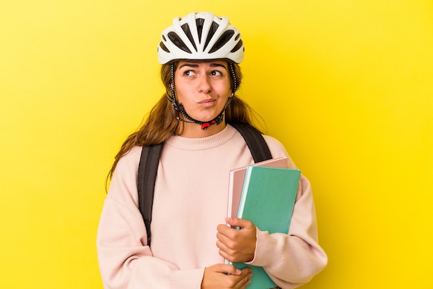 Junge kaukasische Studentin, die einen Fahrradhelm einzeln auf gelbem Hintergrund trägt, verwirrt, fühlt sich zweifelhaft und unsicher.