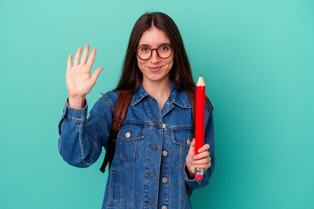 Junge kaukasische Studentin, die einen großen Bleistift auf blauem Hintergrund isoliert hält, lächelt fröhlich und zeigt Nummer fünf mit den Fingern.