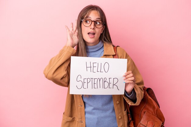 Junge kaukasische Studentin, die hallo September-Plakat auf rosa Hintergrund isoliert hält und versucht, einen Klatsch zu hören.