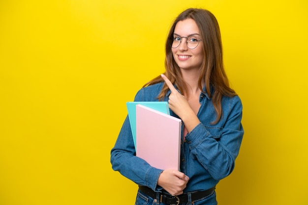 Junge kaukasische Studentin isoliert auf gelbem Hintergrund, die auf die Seite zeigt, um ein Produkt zu präsentieren