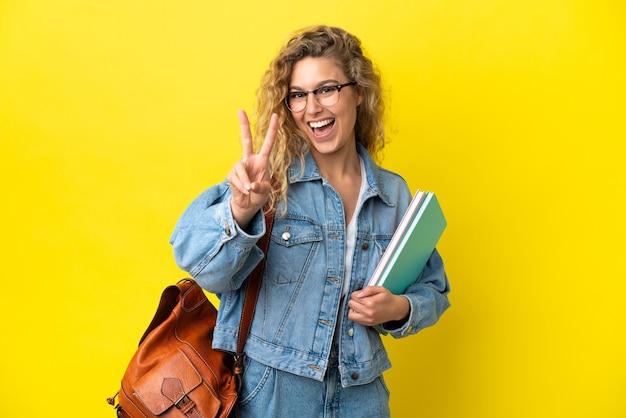 Junge kaukasische Studentin isoliert auf gelbem Hintergrund lächelt und zeigt Victory-Zeichen