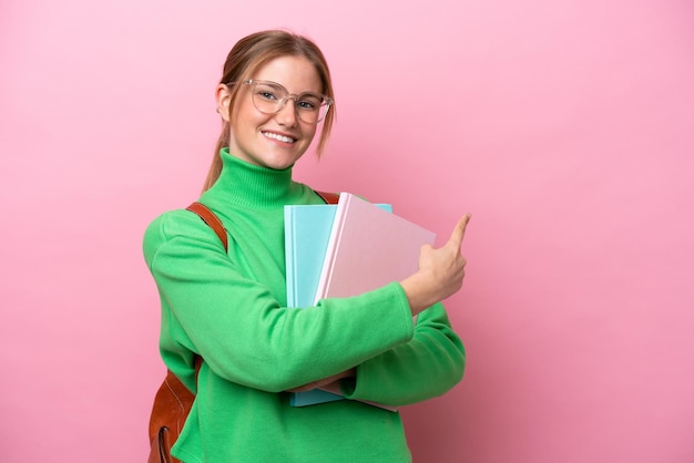 Junge kaukasische Studentin isoliert auf rosafarbenem Hintergrund, die zur Seite zeigt, um ein Produkt zu präsentieren