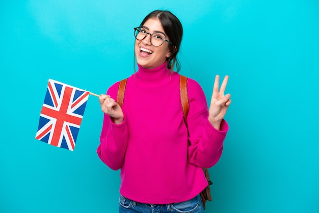 Junge kaukasische Studentin mit englischer Flagge isoliert auf blauem Hintergrund lächelnd und Victory-Zeichen zeigend