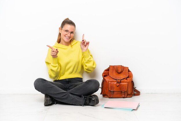 Junge kaukasische Studentin sitzt auf dem Boden isoliert auf weißem Hintergrund und zeigt mit dem Finger auf die Seitenteile und ist glücklich