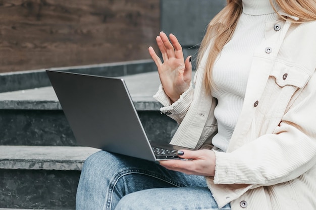 Junge kaukasische Studentin sitzt auf Treppen in der Nähe ihres Colleges mit Laptop, lächelt und hat Vid