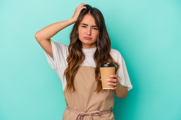 Junge kaukasische Verkäuferin, die einen Kaffee zum Mitnehmen auf blauem Hintergrund hält und schockiert ist, hat sich an ein wichtiges Treffen erinnert.