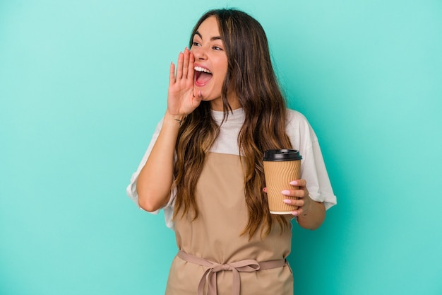 Junge kaukasische Verkäuferin, die einen Kaffee zum Mitnehmen hält, isoliert auf blauem Hintergrund, schreit und hält die Handfläche in der Nähe des geöffneten Mundes.