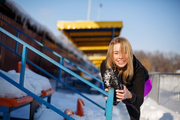 Junge kaukasische weibliche Blondine in den violetten Gamaschen, die Übung auf Tribüne auf einem schneebedeckten Stadion-, Sitz- und Sportlebensstil ausdehnen