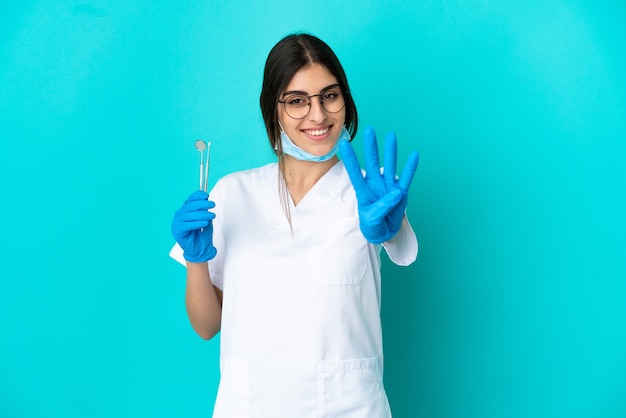 Junge kaukasische Zahnarztfrau, die Werkzeuge lokalisiert auf blauem Hintergrund glücklich hält und vier mit den Fingern zählt