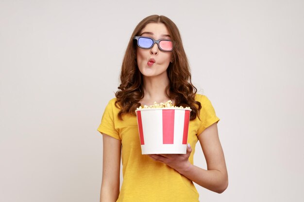 Junge kindliche Frau in 3D-Brille und gelbem lässigem T-Shirt, die einen Filmfilm mit einem Eimer Popcorn anschaut, der Fischgesichtsgrimasse mit Schmolllippen macht Studioaufnahme isoliert auf grauem Hintergrund