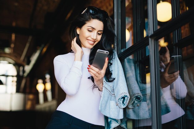 Junge, kleinliche, brünette Geschäftsfrau in eleganter Kleidung, die vor dem Café-Interieur steht und darauf wartet, dass sich die Leute treffen, indem sie drinnen mit dem Smartphone SMS-Nachrichten online senden Kommunikationskonzept