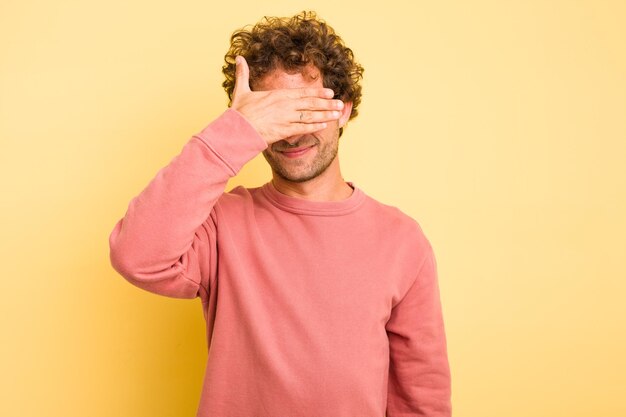 Junge, kluge, kaukasische Mann auf gelbem Hintergrund bedeckt die Augen mit Händen und lächelt