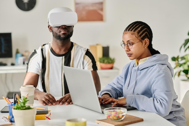 Junge Kollegen nutzen Laptop, um VR-Brillen zu testen, während sie am Tisch im Büro sitzen