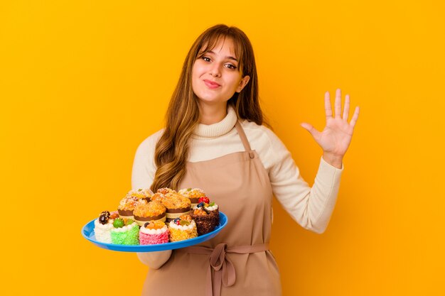 Junge Konditorfrau, die auf gelbem Hintergrund lokalisiert wird, lächelt fröhlich und zeigt Nummer fünf mit den Fingern.