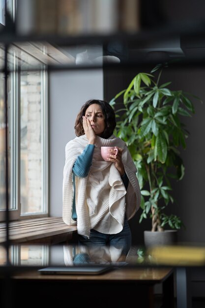 Junge kranke ungesunde weibliche Angestellte mit saisonalen Grippesymptomen, die heißen Tee im Büro trinken