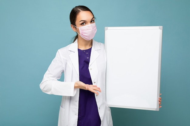 Junge Krankenschwester in der schützenden Gesichtsmaske und im weißen medizinischen Mantel, die eine leere Magnettafel lokalisiert halten