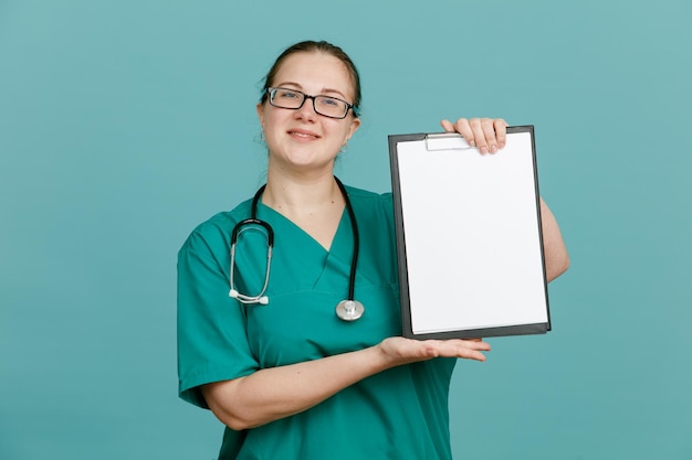 Junge krankenschwester in medizinischer uniform mit stethoskop um den hals, die ein klemmbrett hält und in die kamera blickt, glücklich und positiv lächelnd, selbstbewusst vor blauem hintergrund