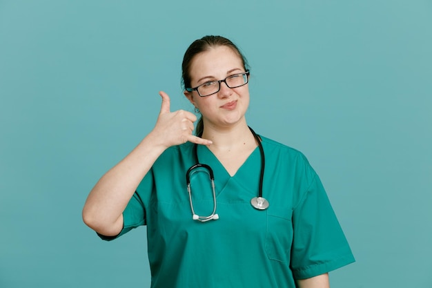 Junge Krankenschwester in medizinischer Uniform mit Stethoskop um den Hals, die glücklich und positiv in die Kamera schaut und mich mit einer Geste anruft, die über blauem Hintergrund steht