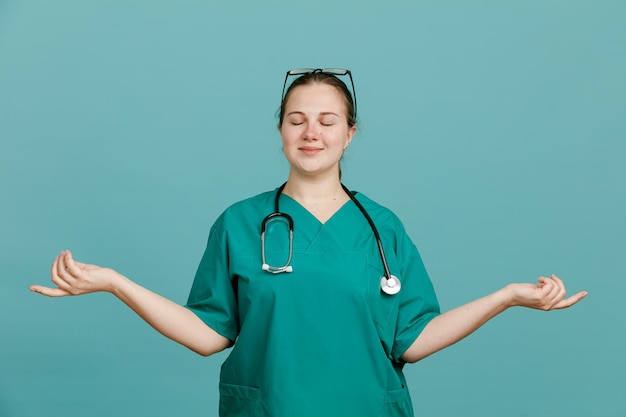 Junge Krankenschwester in medizinischer Uniform mit Stethoskop um den Hals, die ruhig und entspannt aussieht und Meditationsgesten mit über blauem Hintergrund stehenden Händen macht