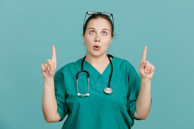 Junge Krankenschwester in medizinischer Uniform mit Stethoskop um den Hals, die überrascht nach oben zeigt und mit dem Zeigefinger nach oben zeigt, der über blauem Hintergrund steht