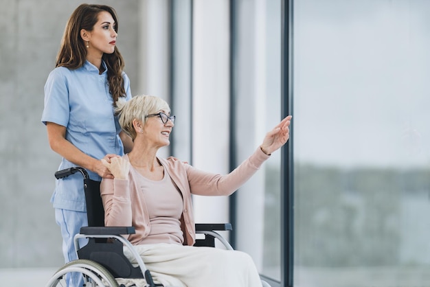 Junge Krankenschwester und Seniorin im Rollstuhl, die durch das Fenster eines Pflegeheims schauen.