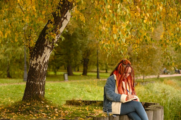 Junge kreative Frau mit Dreadlocks, die das Schreiben in ein Notizbuch zeichnet, das auf Baumstümpfen in der Nähe eines Sees in einer Parklandschaft sitzt