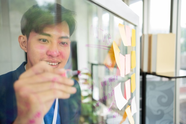 Junge kreative Geschäftsmann hält einen Marker und schreibt einen Plan und teilt seine Idee an einer Glaswand mit einer klebrigen Notiz Brainstorming und Diskussion und Formulierung von Geschäftsstrategien im Tech-Startup-Büro