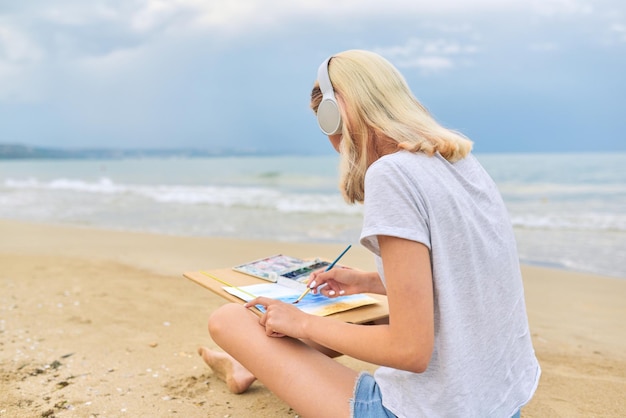 Junge Künstlerin malt mit Wasserfarben an der Küste sitzend, kreative Frau in Kopfhörern, die Musik hört, schöner Meereslandschaftshintergrund, Kopierraum