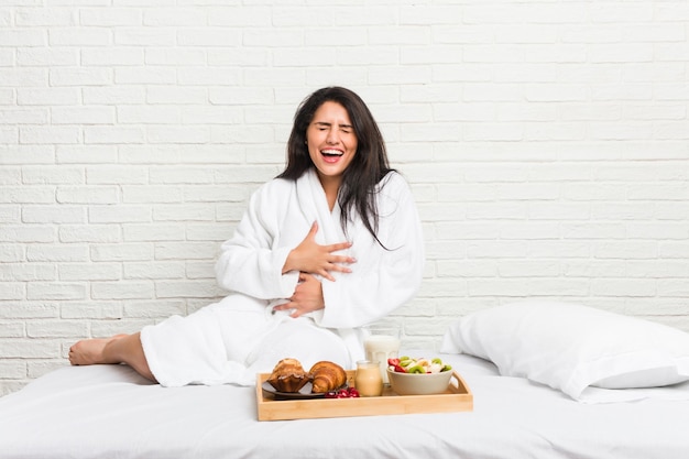 Junge kurvige Frau, die ein Frühstück auf dem Bett nimmt, lacht glücklich und hat Spaß, Hände auf Bauch zu halten.