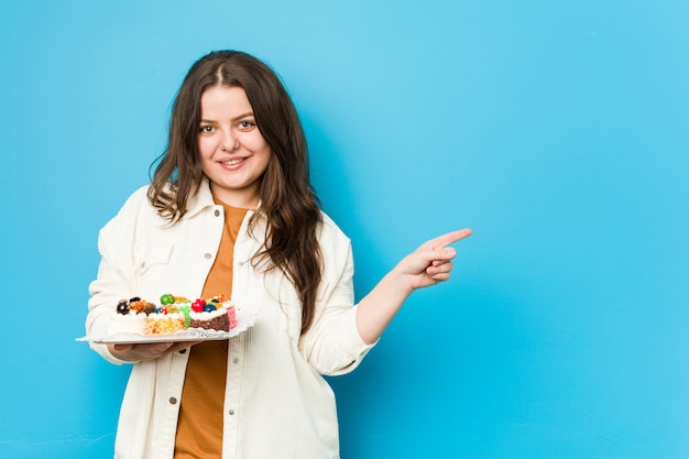 Junge kurvige Frau, die einen süßen Kuchen hält, der lächelt und zur Seite zeigt und etwas an der leeren Stelle zeigt.