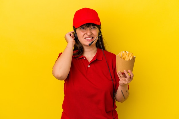 Junge kurvige kaukasische Frau Fast-Food-Restaurant-Arbeiter, die Pommes auf blauem Hintergrund isoliert hält und die Ohren mit den Händen bedeckt.