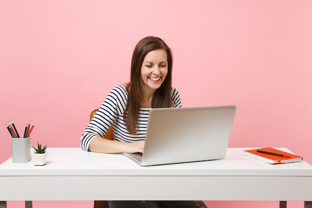 Junge lachende Frau in Freizeitkleidung, die mit Laptop an einem Projekt arbeitet, während sie im Büro sitzt