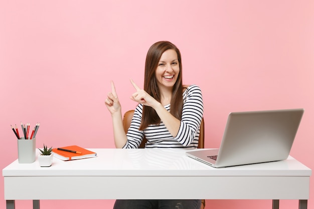 Junge lachende Frau in Freizeitkleidung zeigen mit dem Zeigefinger beiseite sitzen, arbeiten am weißen Schreibtisch mit modernem PC-Laptop