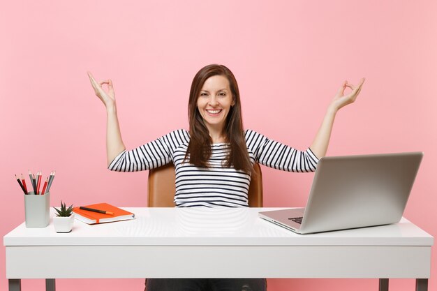 Junge lachende Frauenmeditation verteilt Hände in Yoga-Pose sitzen nach der Arbeit am weißen Schreibtisch mit zeitgenössischem PC-Laptop einzeln auf pastellrosa Hintergrund. Erfolgsgeschäftskarrierekonzept. Platz kopieren.