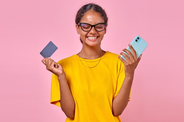 Junge, lachende indische Frau mit Telefon und Kreditkarte steht in einem rosa Studio