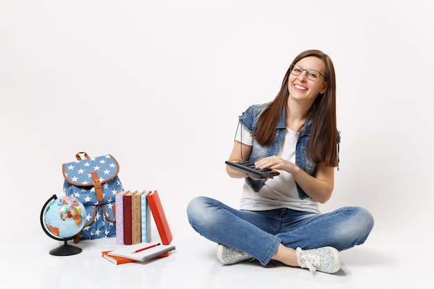 Junge lachende intelligente Studentin, die einen Taschenrechner hält und verwendet, um mathematische Gleichungen zu lösen, die in der Nähe von Globus, Rucksack, Schulbüchern isoliert sitzen