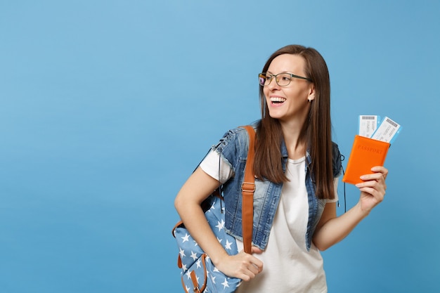 Junge lachende Studentin in Gläsern mit Rucksack, die beiseite schaut, halten Pass, Bordkarten einzeln auf blauem Hintergrund. Ausbildung an einer Hochschule im Ausland. Flugreise-Flugkonzept.