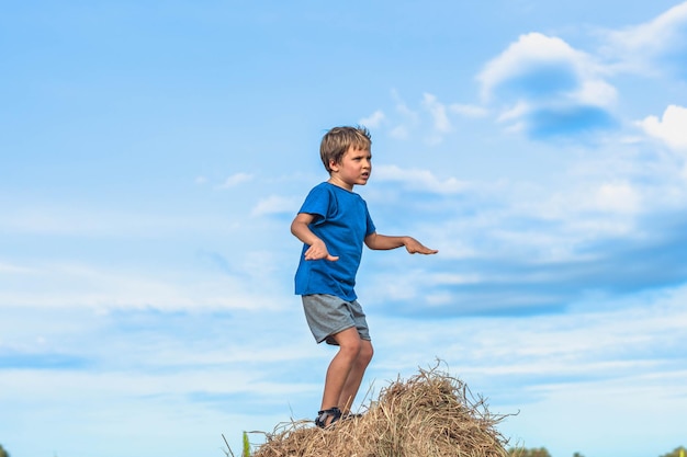 Junge Lächeln spielen Tanz Grimasse zeigen blaues T-Shirt stehen auf Heuhaufen Ballen aus trockenem Gras klarer Himmel sonniger Tag Gleichgewichtstraining Konzept glückliche Kindheit Kinder im Freien saubere Luft in der Nähe der Natur