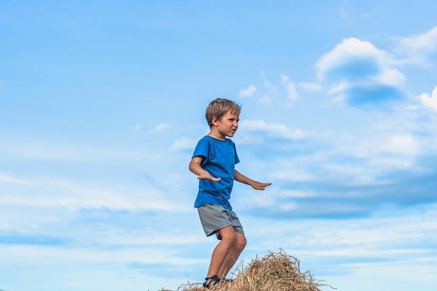 Junge lächeln spielen tanz grimasse zeigen blaues t-shirt stehen auf heuhaufen ballen aus trockenem gras klarer himmel sonniger tag gleichgewichtstraining konzept glückliche kindheit kinder im freien saubere luft in der nähe der natur