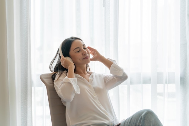 Junge lächelnde asiatische Frau, die sitzt und Musik hört Junges Brunettemädchen, das zu Hause in einem Freizeit timex9Copy Raum sich entspannt